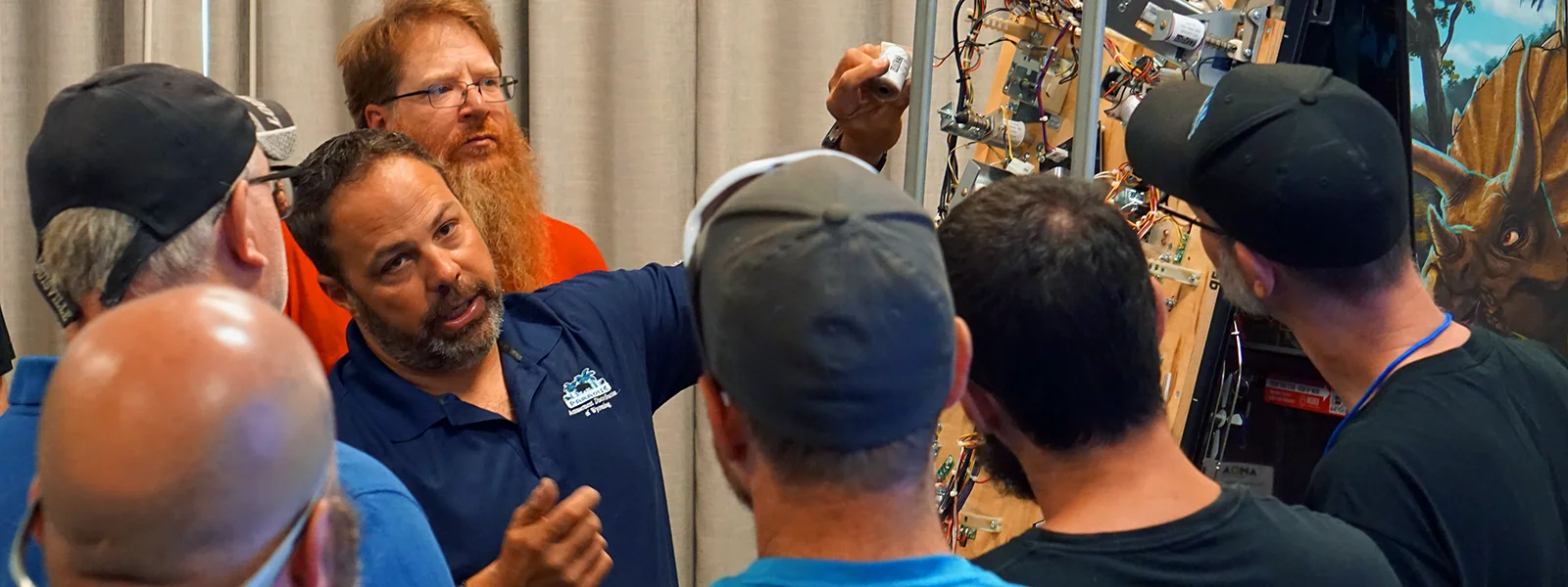 A photo of students gathering around a lecturer demonstrating principles on a disassembled pinball machine
